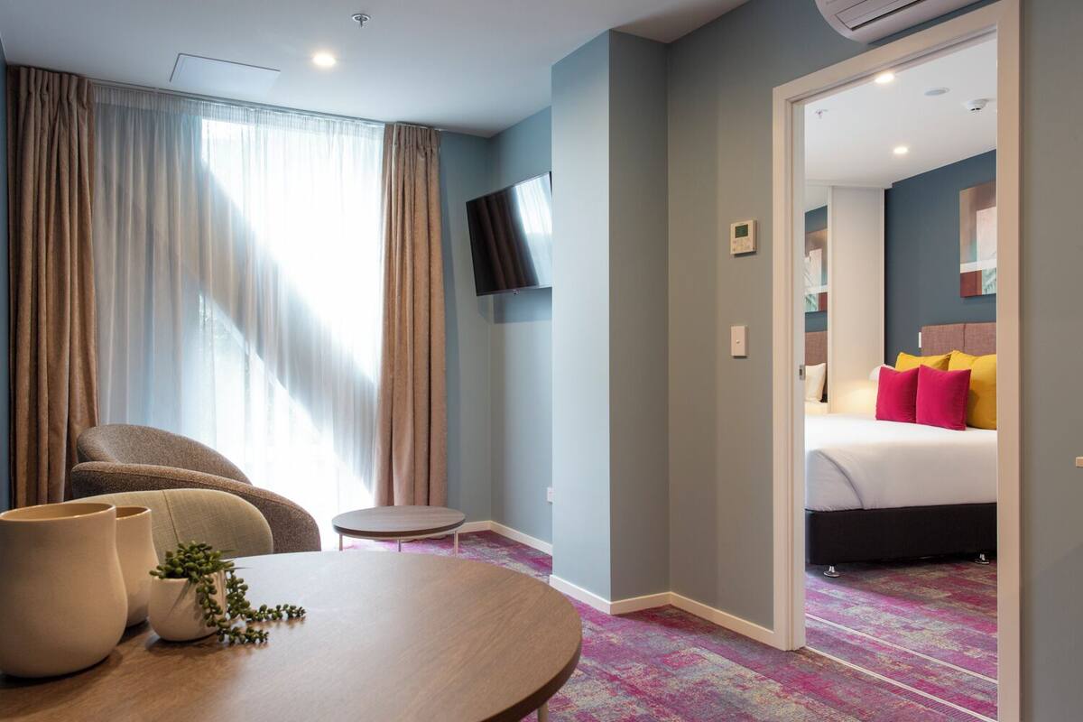 A hotel room with a bed, a TV mounted on the wall, a small round table, and a potted plant.