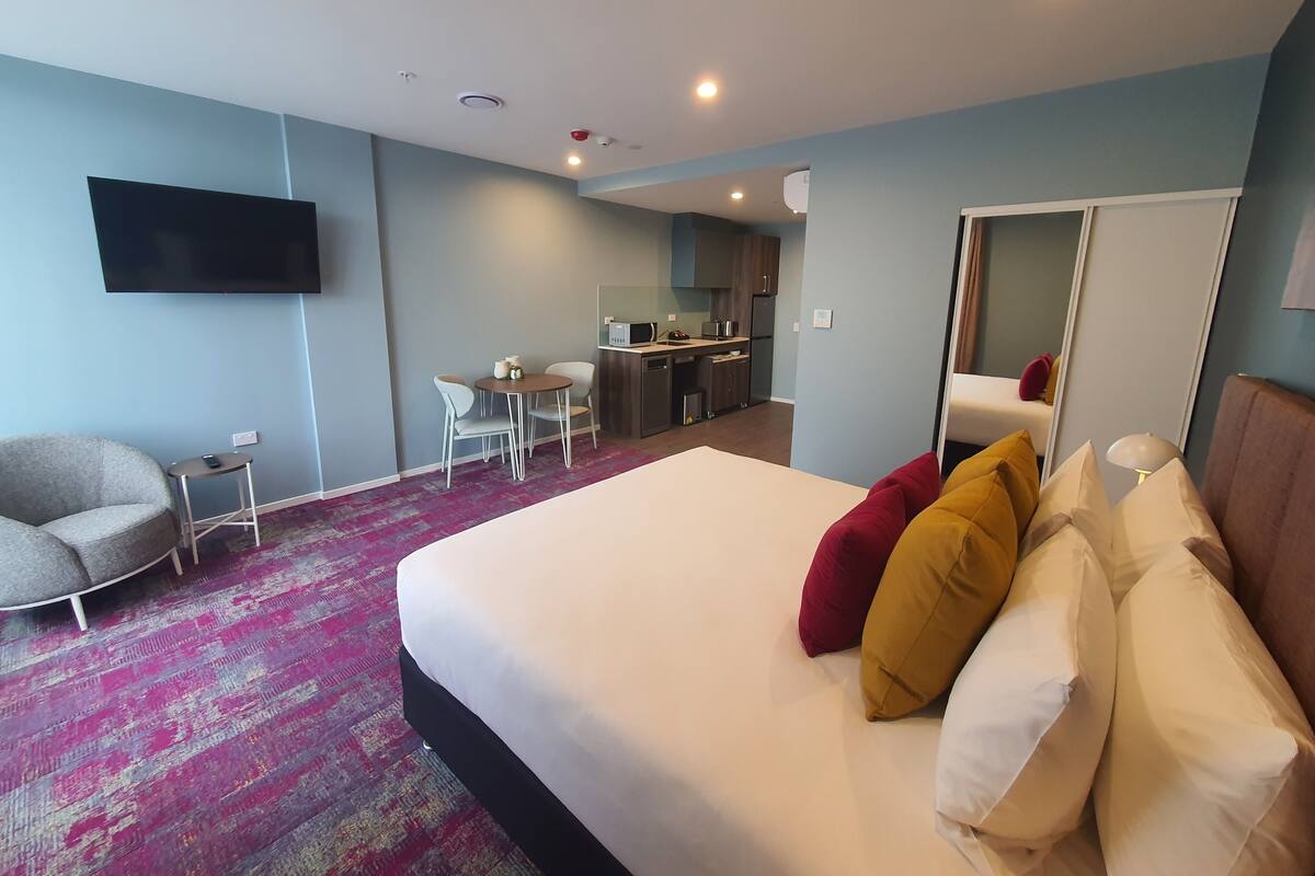 A hotel room with a bed, a desk, a chair, a TV, and a kitchenette.