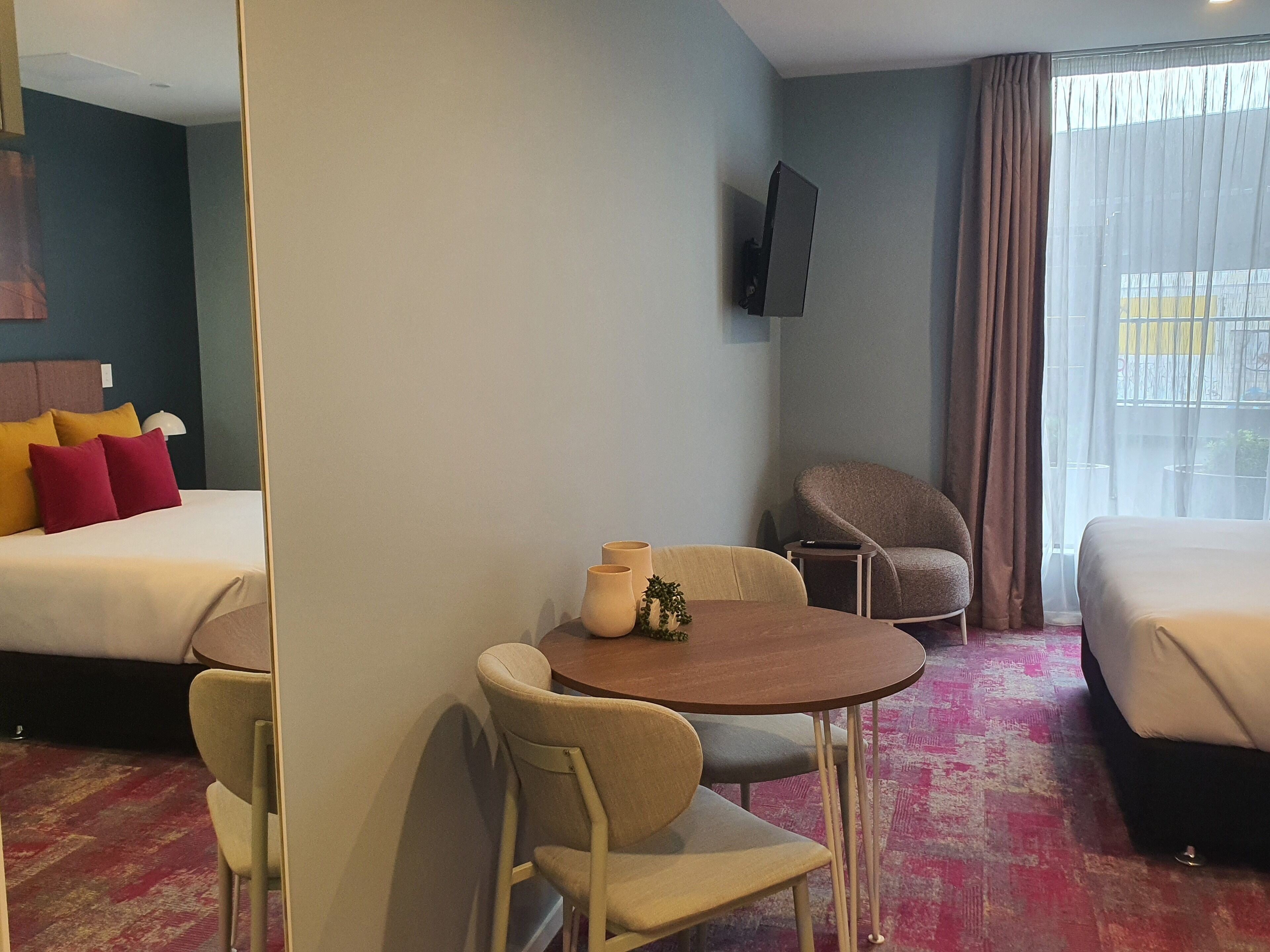 A hotel room with two beds, a dining table with chairs, a TV mounted on the wall, and a large window with curtains.