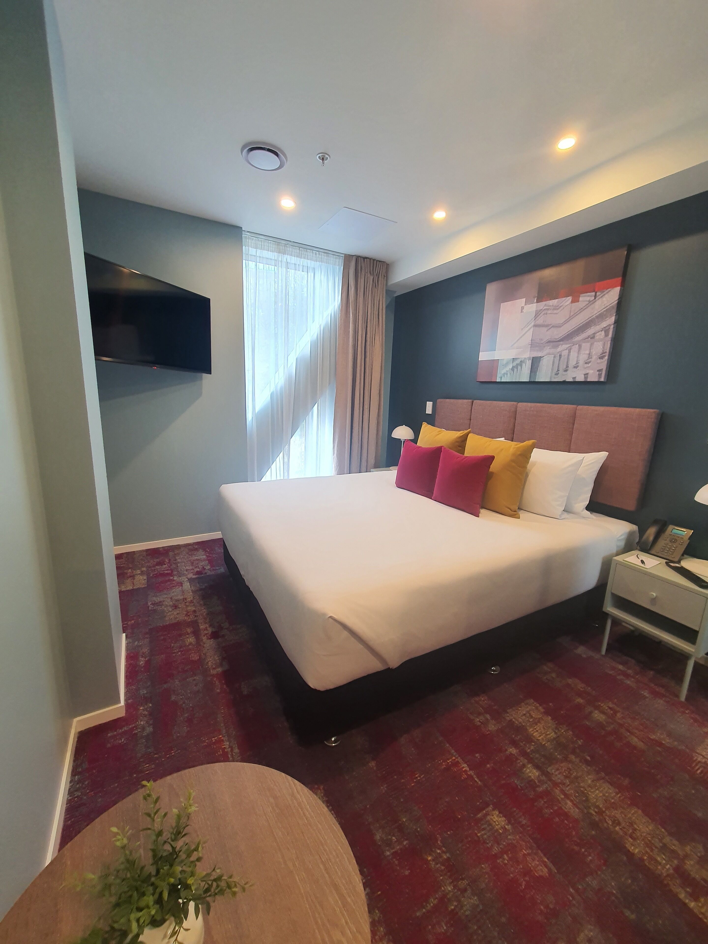A hotel room with a large bed, a flat-screen TV mounted on the wall, a bedside table with a phone, and a window with curtains.