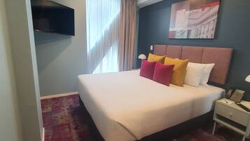 A hotel room with a large bed, a flat-screen TV mounted on the wall, a bedside table with a phone, and a window with curtains.
