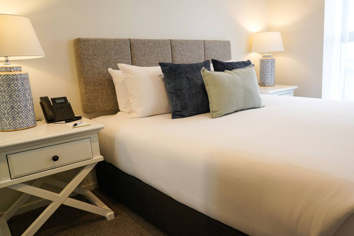 A neatly made bed with white and dark pillows, a grey headboard, and two bedside tables with lamps and a phone.