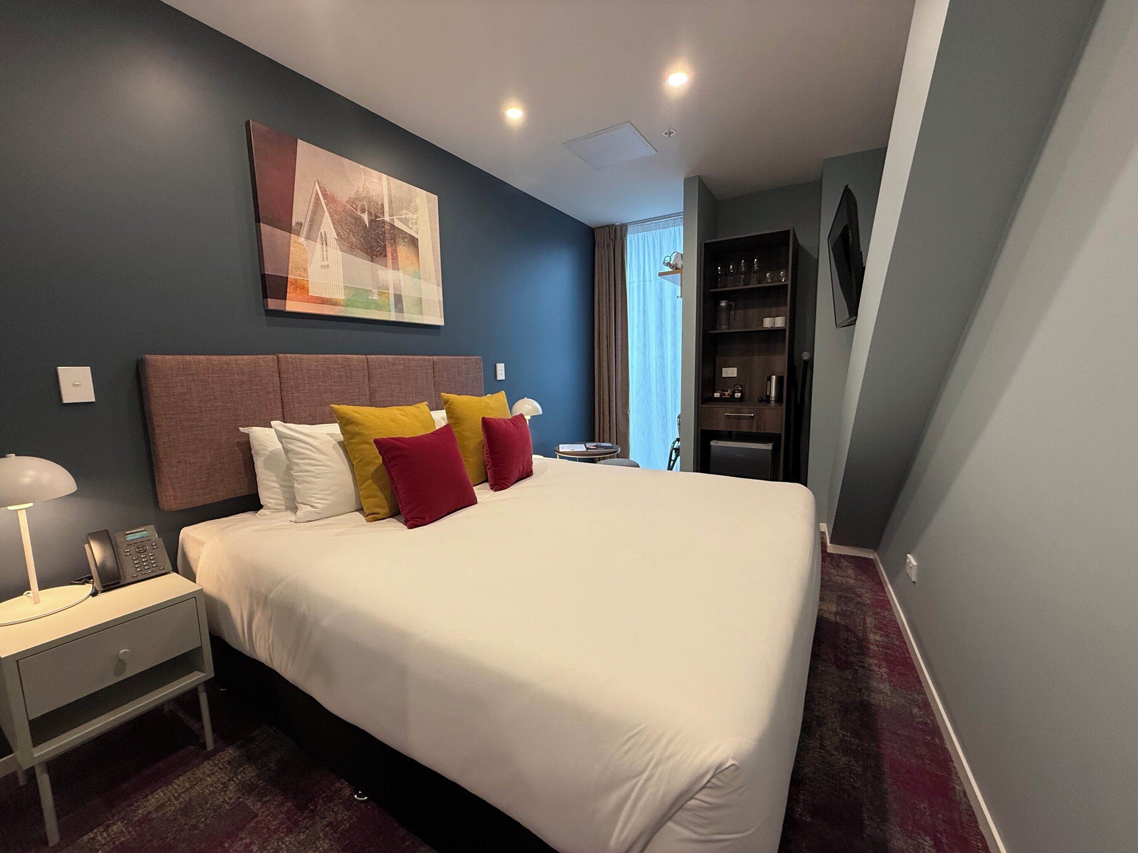 A hotel room with a large bed, bedside table, lamp, and a painting on the wall.
