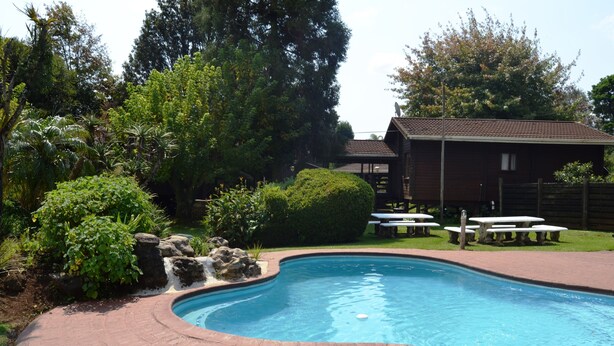 A swimming pool with a brick border, surrounded by greenery and a wooden building with picnic tables. - Log Cabin & Settlers Village (Graskop)