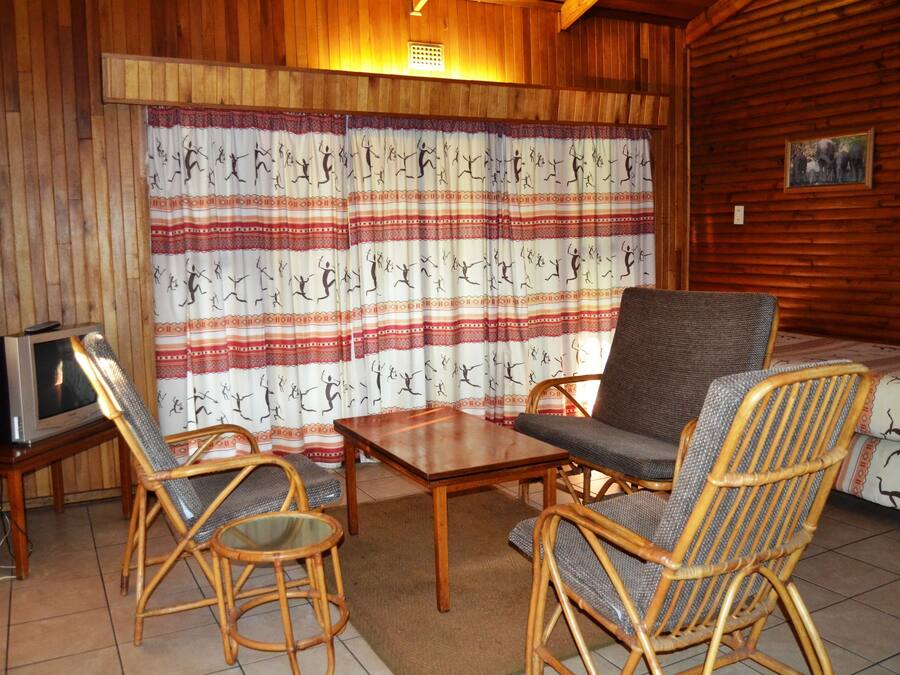 Una habitación acogedora con paredes de madera, un televisor sobre un mueble de madera, dos sillas de mimbre, una mesita de madera y una cortina estampada.
