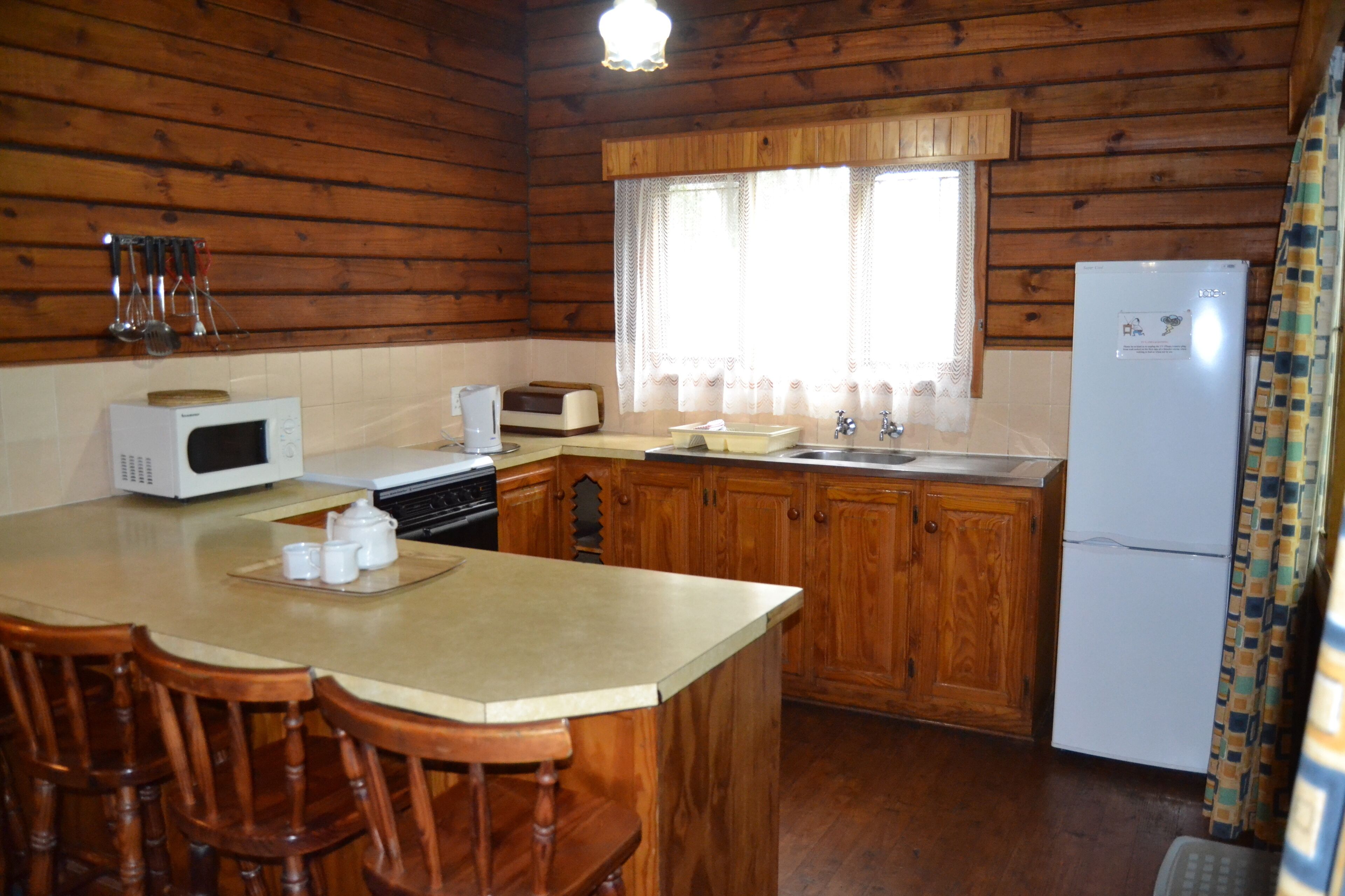 Cocina de cabaña de madera con microondas, tostadora, lavabo y refrigerador. Hay sillas de madera en la isla de la cocina.