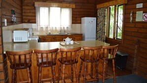 A wooden cabin kitchen with a central island, bar stools, a refrigerator, and a window with curtains. - Log Cabin & Settlers Village (Graskop)