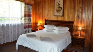 A wooden-paneled room with a large bed, two bedside lamps, and a window with sheer curtains. - Log Cabin & Settlers Village (Graskop)