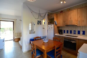 A compact kitchen with wooden cabinets, a dining table with blue cushioned chairs, a refrigerator, and a coffee maker.