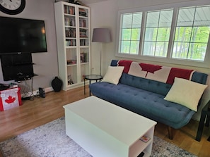 Ein Wohnzimmer mit blauem Sofa, weißem Couchtisch, Bücherregal, Fernseher und einem Fenster mit Blick auf Bäume.