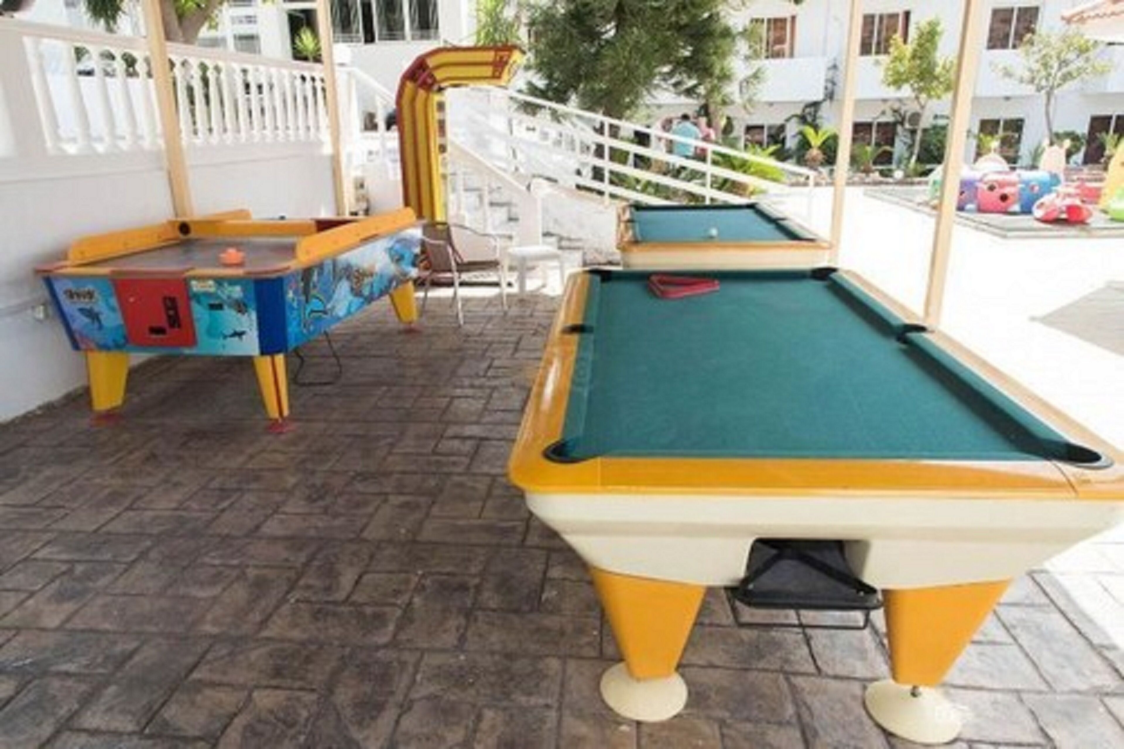 Une table de billard recouverte d’un drap vert, avec des pieds jaunes, placée à l’extérieur sur une surface pavée, avec une clôture blanche et un escalier en arrière-plan.