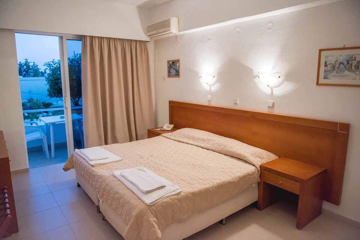 A hotel room with a bed, a bedside table, a nightstand, a window with curtains, an air conditioning unit, and a small table with chairs outside.