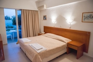 Ein Hotelzimmer mit Bett, Nachttisch, Klimaanlage und einem kleinen Tisch mit Stühlen davor. Ein Fenster mit Vorhängen ist ebenfalls vorhanden.