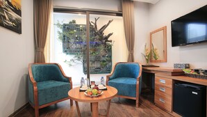 A compact hotel room with two teal chairs, a wooden table with fruit and drinks, a small kitchenette, a TV, and a large window with a view of greenery. - Oriana Hotel (Hanoi)