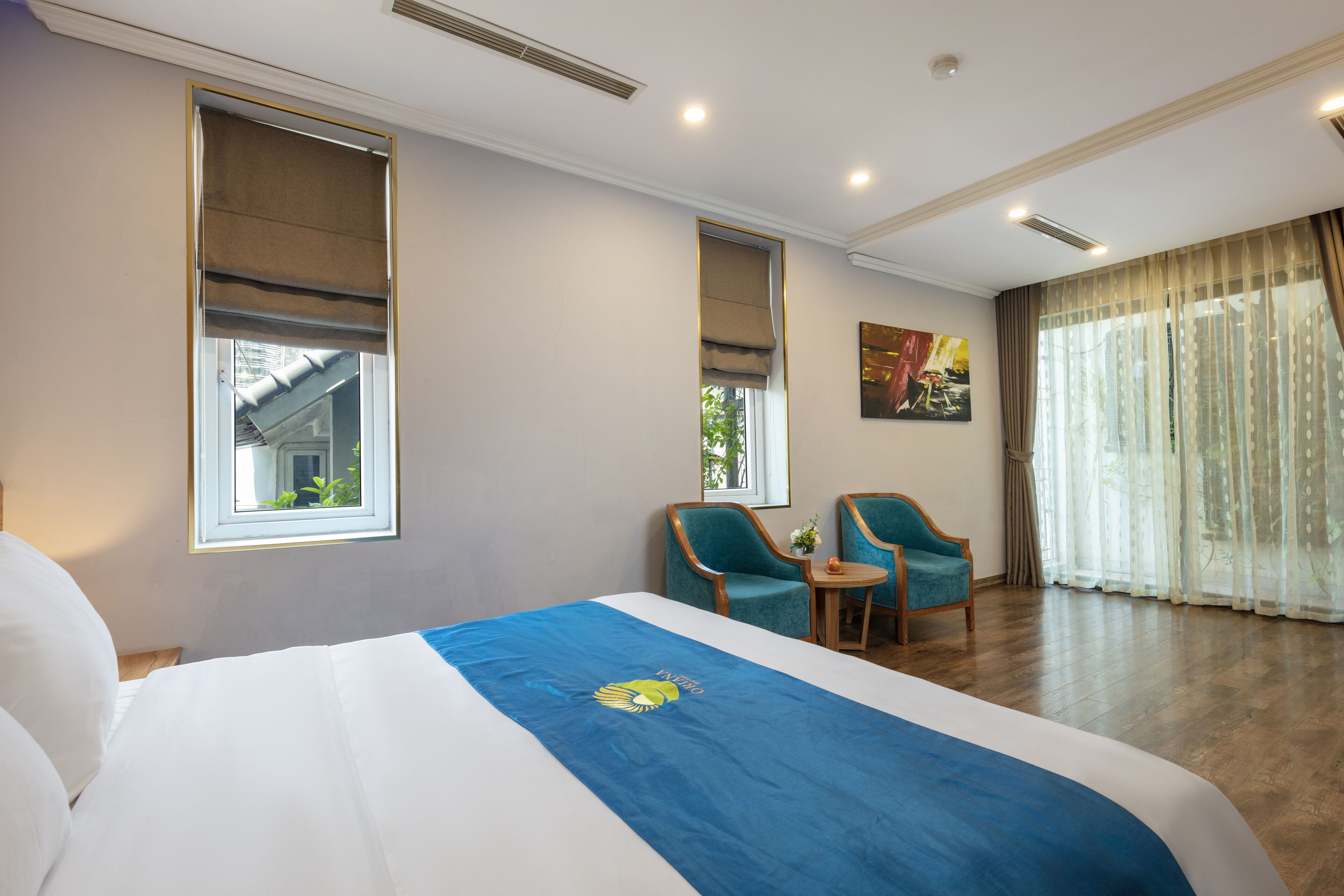 A hotel room with a large bed, two armchairs, a small table, and a view of the outdoors through the windows.