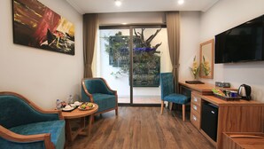 A modern room with a wooden floor, teal chairs, a small table with a plate of fruit, a TV, and a large window with a view of greenery. - Oriana Hotel (Hanoi)