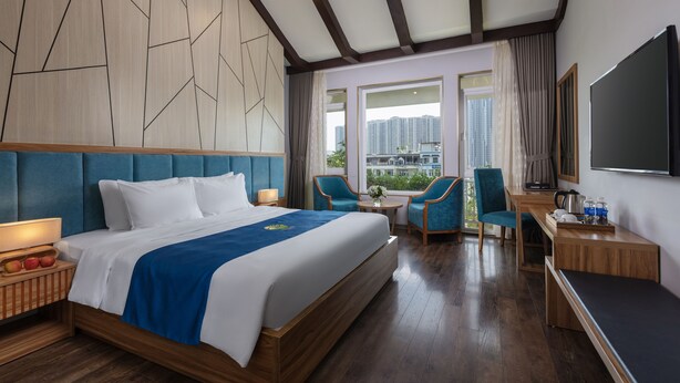A hotel room with a large bed, a desk, two chairs, a TV, and a view of the city. - Oriana Hotel (Hanoi)