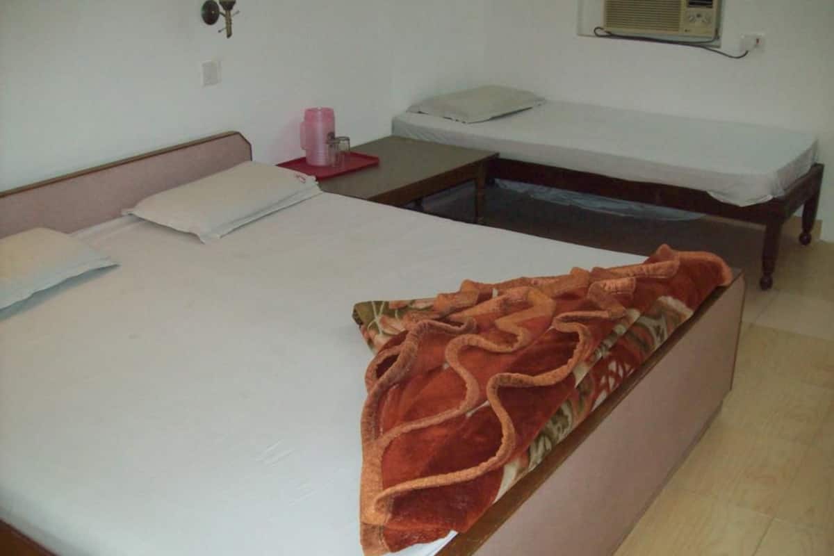 A room with two beds, one with a red blanket and the other with a white sheet. There is a small table with a pink object on it, and a wall-mounted heater.