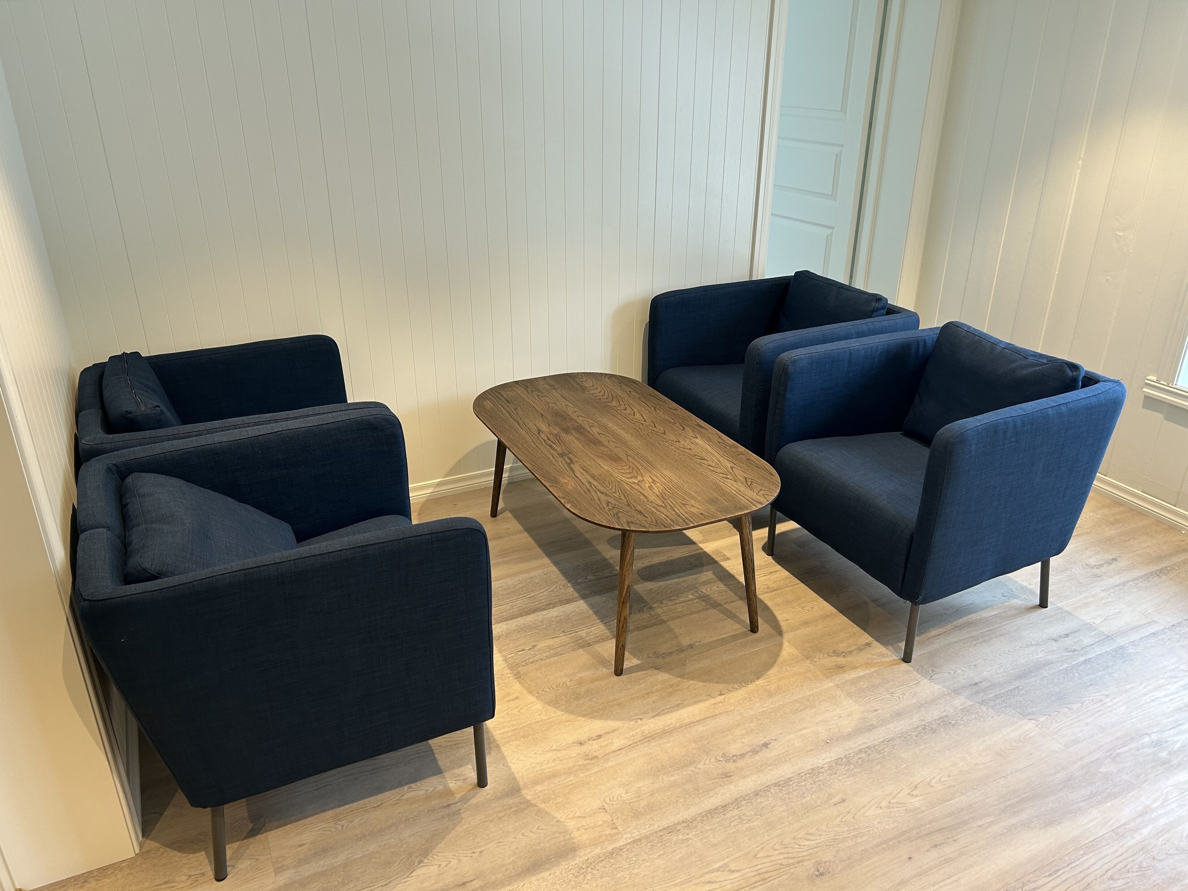 Une pièce dotée d’un plancher en bois, de murs recouverts de lambris blancs et de trois fauteuils bleu foncé disposés autour d’une table basse centrale en bois.