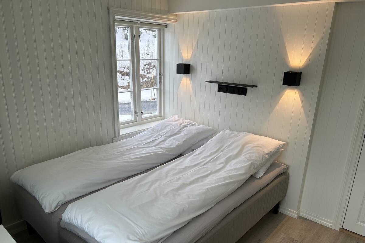 A single bed with white bedding in a room with white paneled walls and a window.