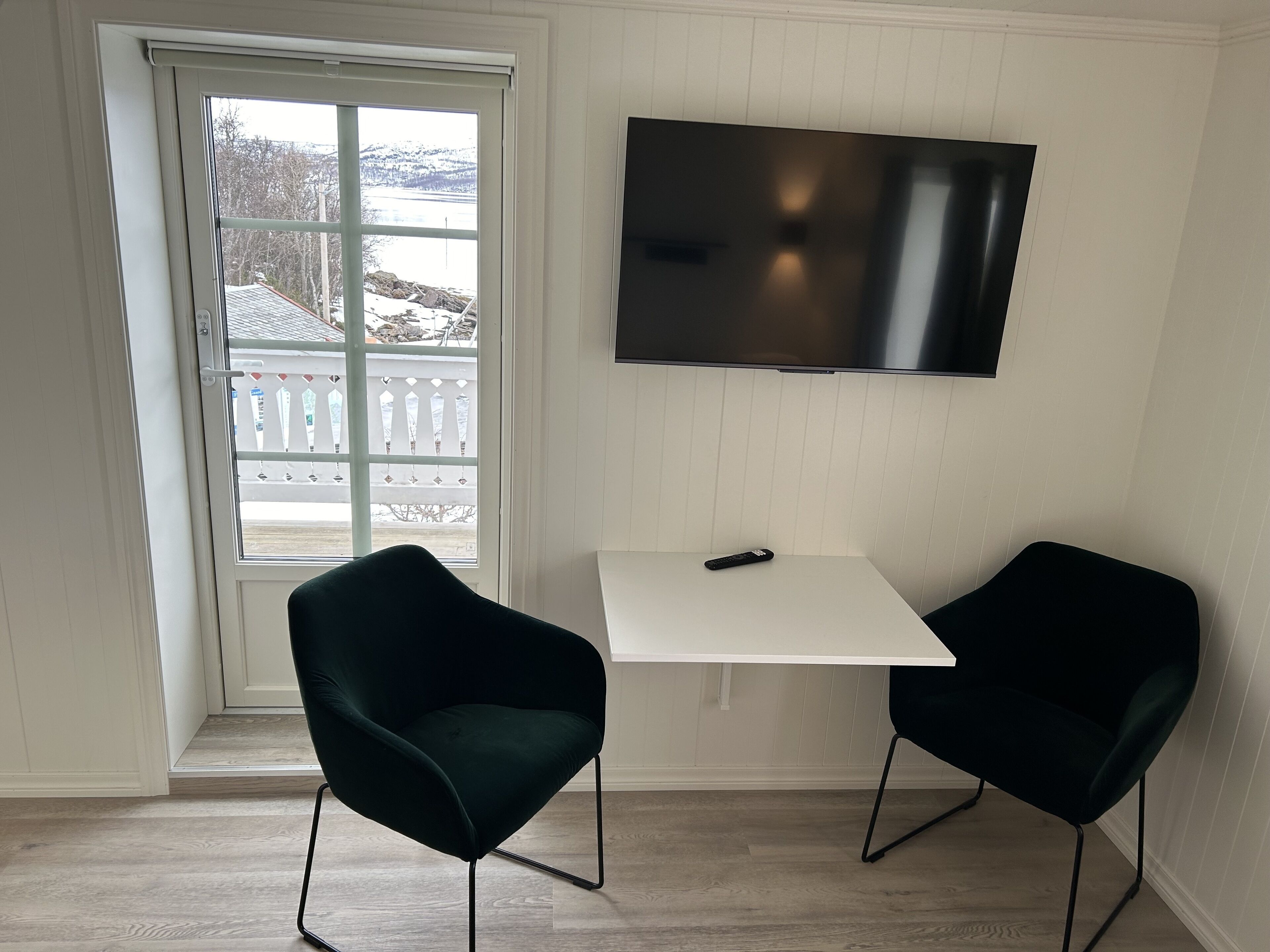 A room with a white table and two black chairs, a TV mounted on the wall, and a window with a view of a snowy landscape.