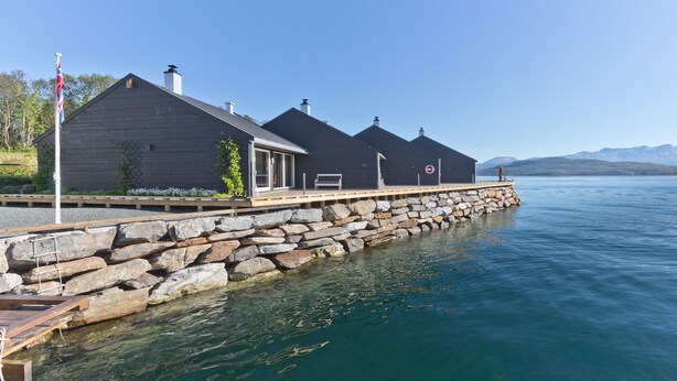 A waterfront property with a stone pier, a wooden deck, and a row of dark-colored buildings. - Finsnes Gaard (Senja)