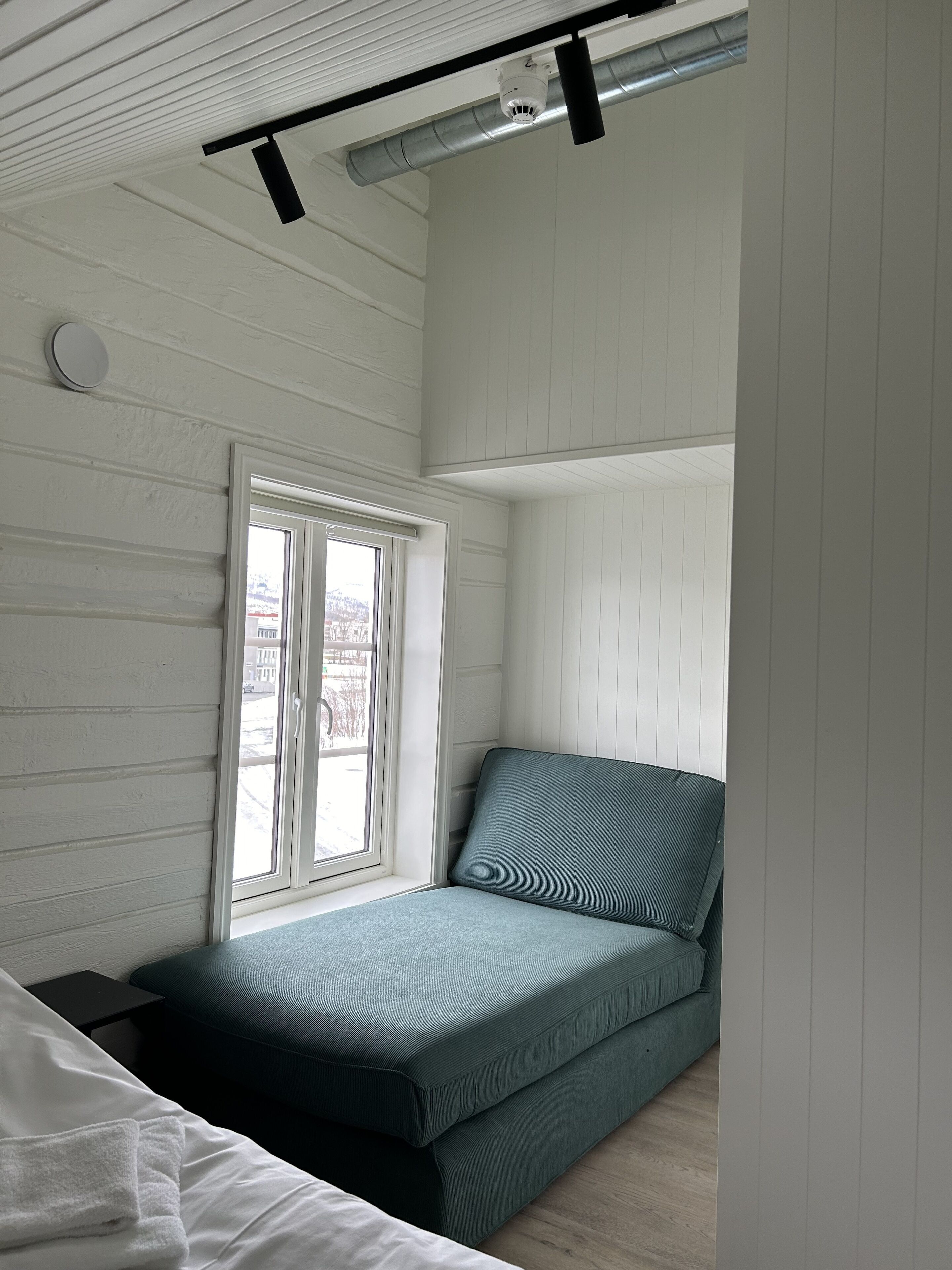 Una habitación con una cama, una ventana con persianas, un respiradero en el techo y una lámpara fijada a la pared.