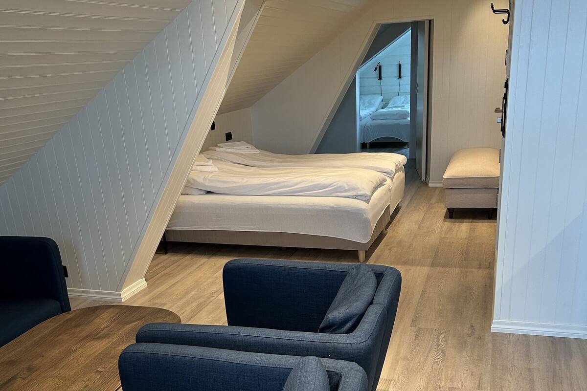 A modern, compact room with a lofted bed, a small seating area with sofas, and a wooden floor.