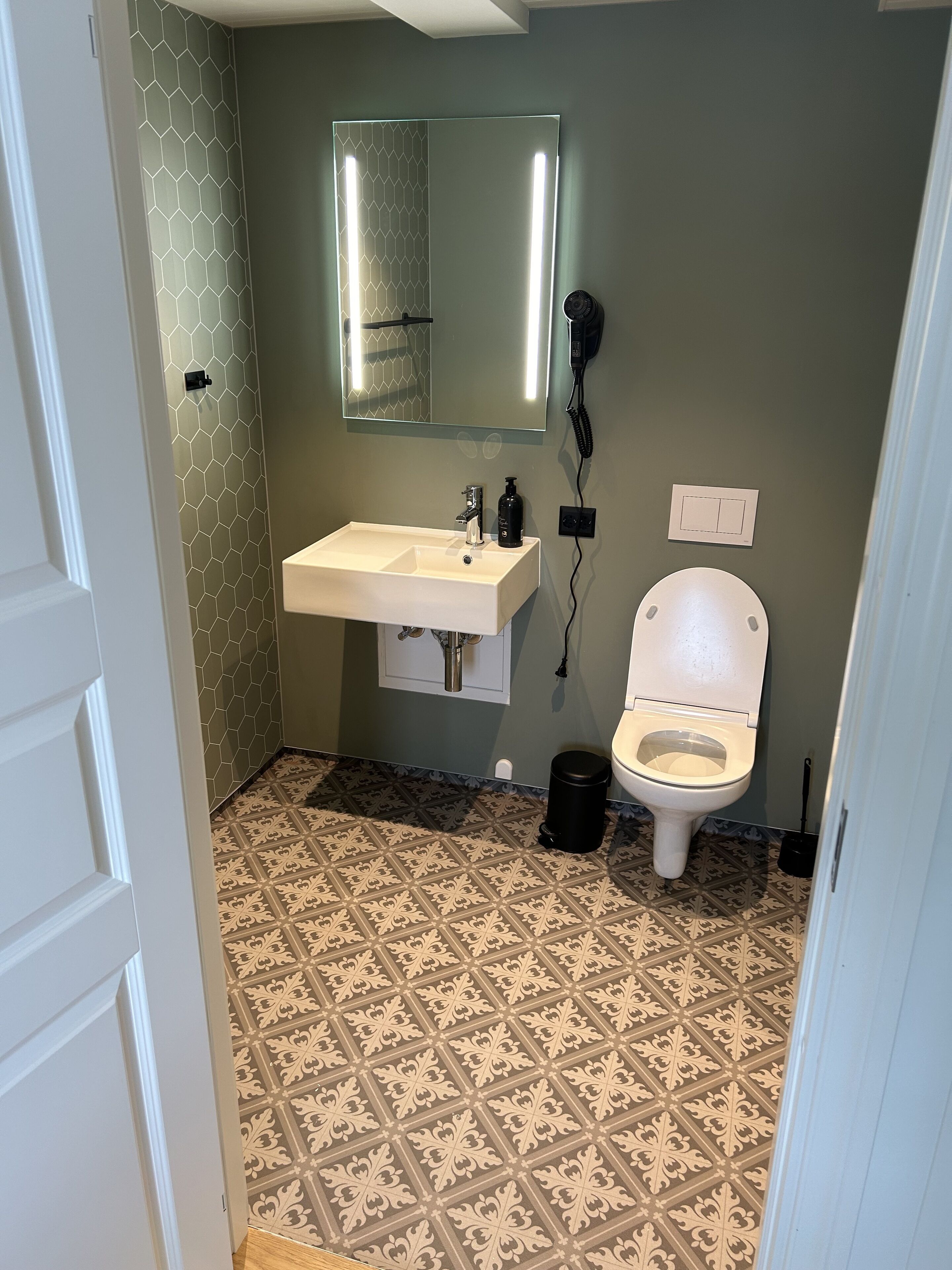 A bathroom with a white sink, a mirror, a toilet, and patterned floor tiles.