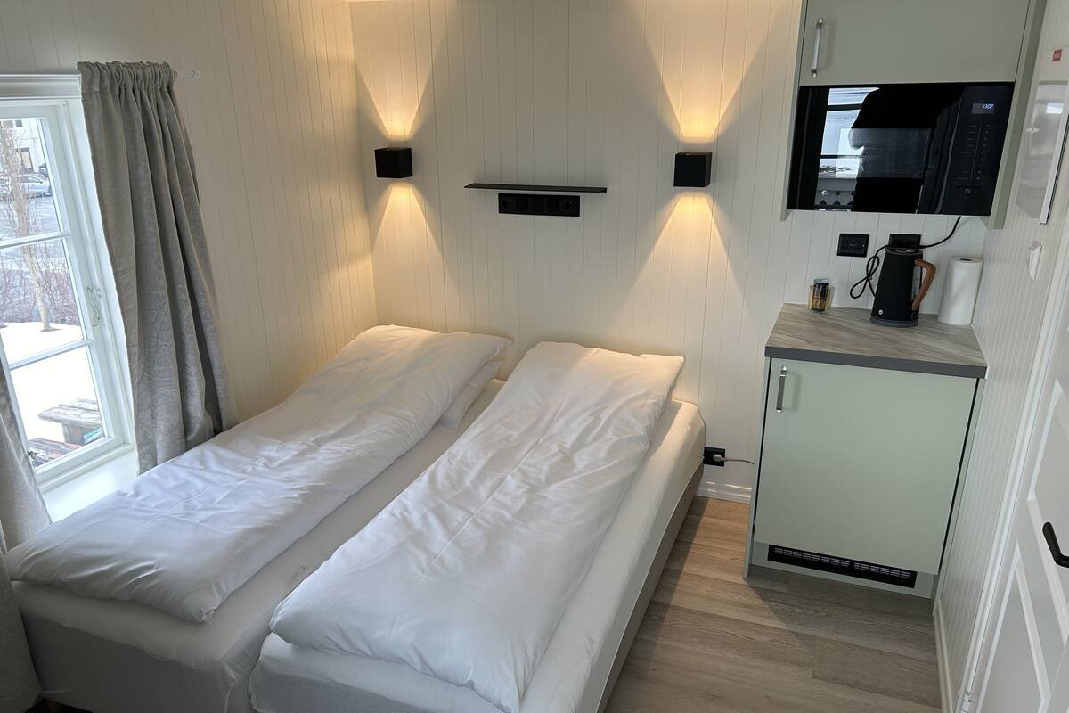 A room with two single beds, a small kitchenette, a window with curtains, and a wall-mounted TV.