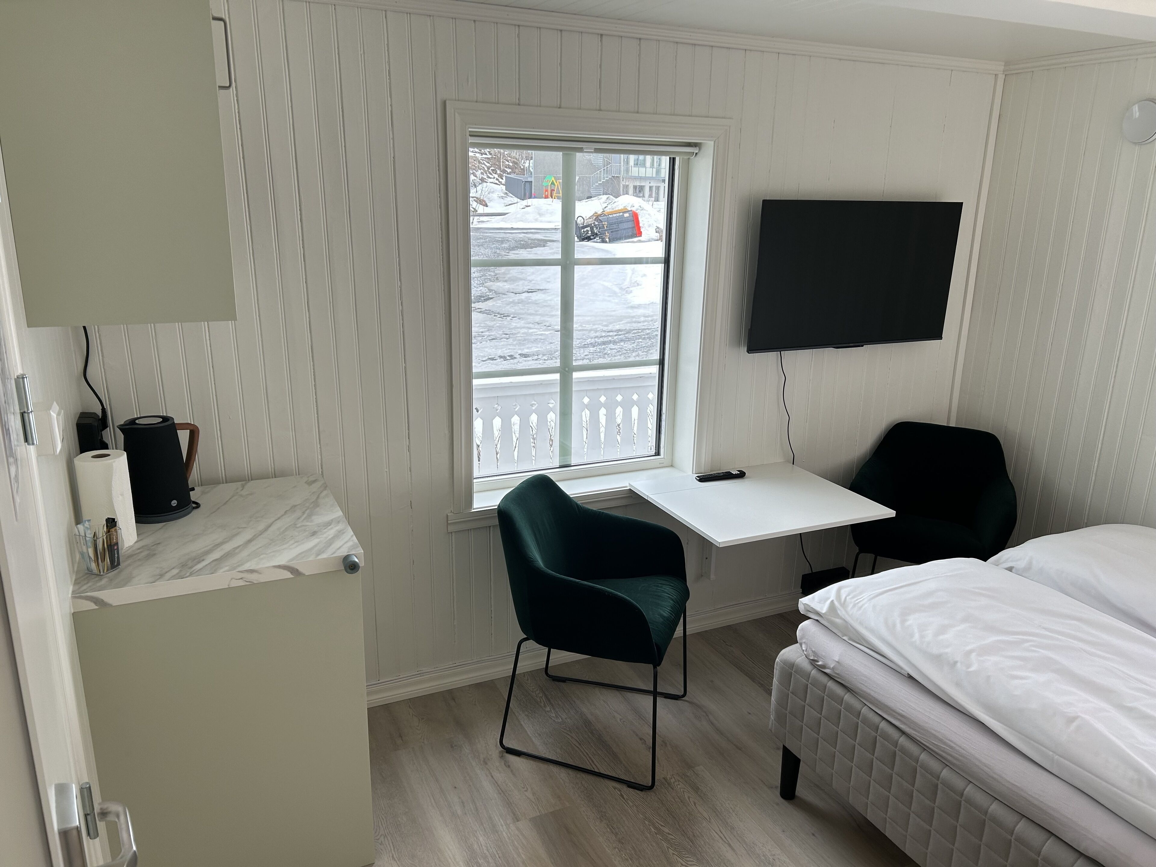 A hotel room with a bed, a desk with a chair, a TV mounted on the wall, and a window with a view of the outside.