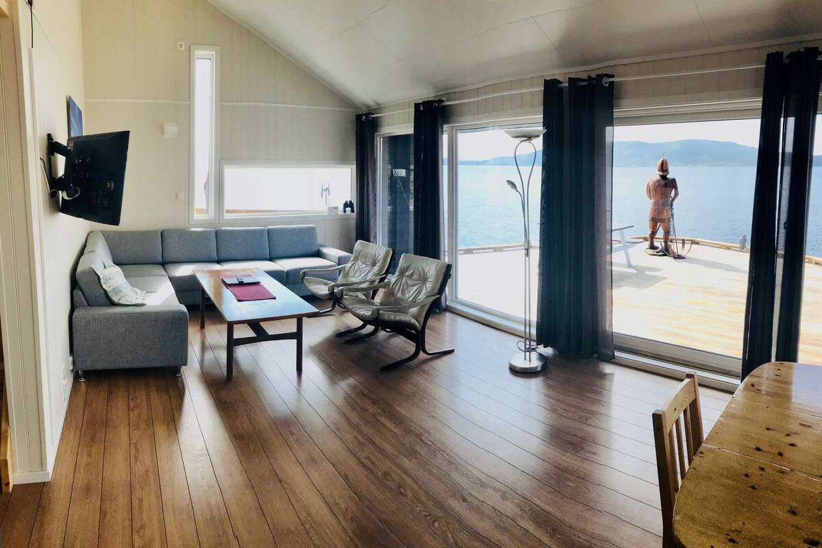 A living room with a grey sofa, a wooden coffee table, and two leather chairs. There is a large window with a view of the sea and mountains.