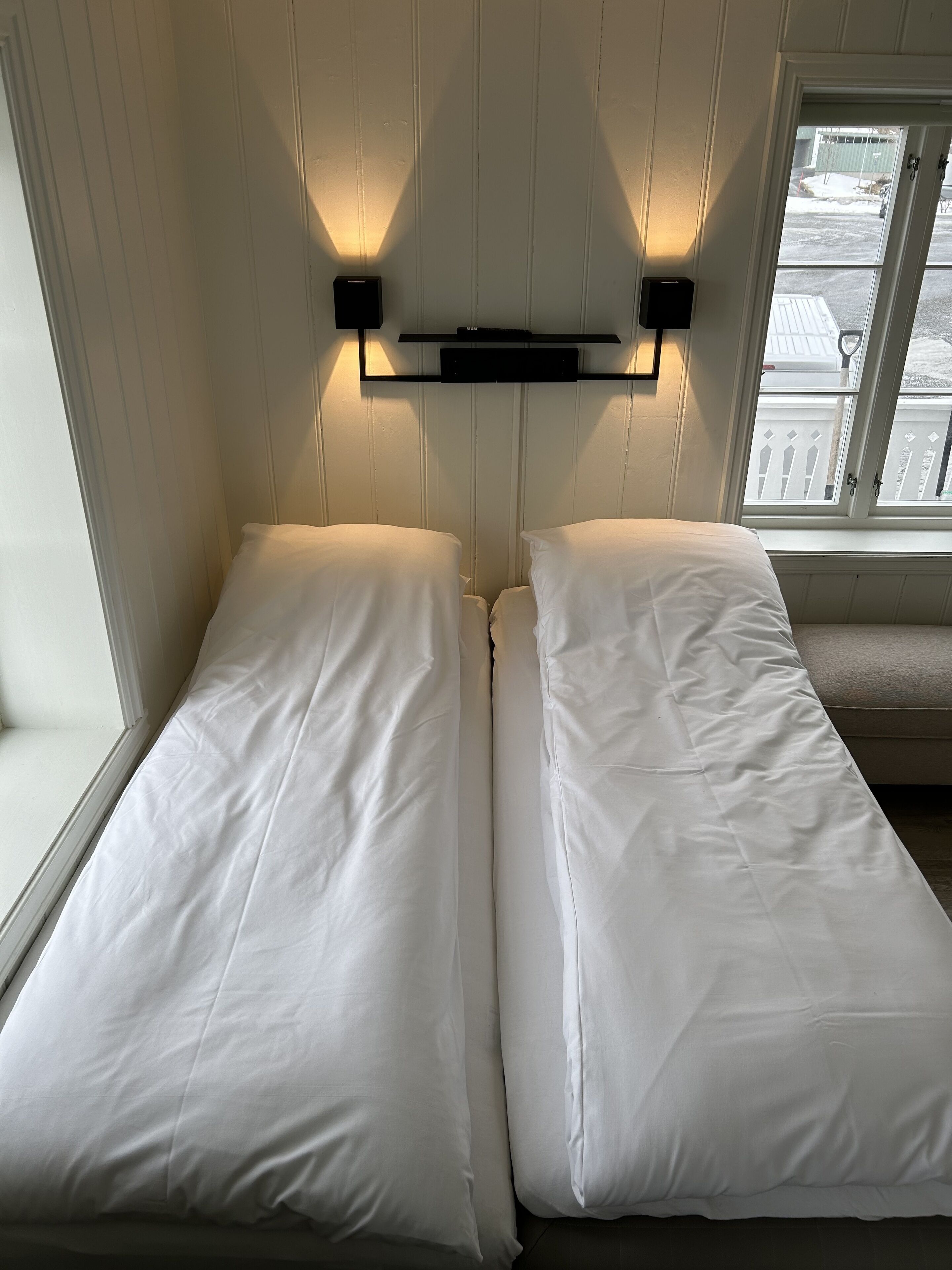 A room with two single beds, a window with a view of the outside, and wall-mounted lighting.