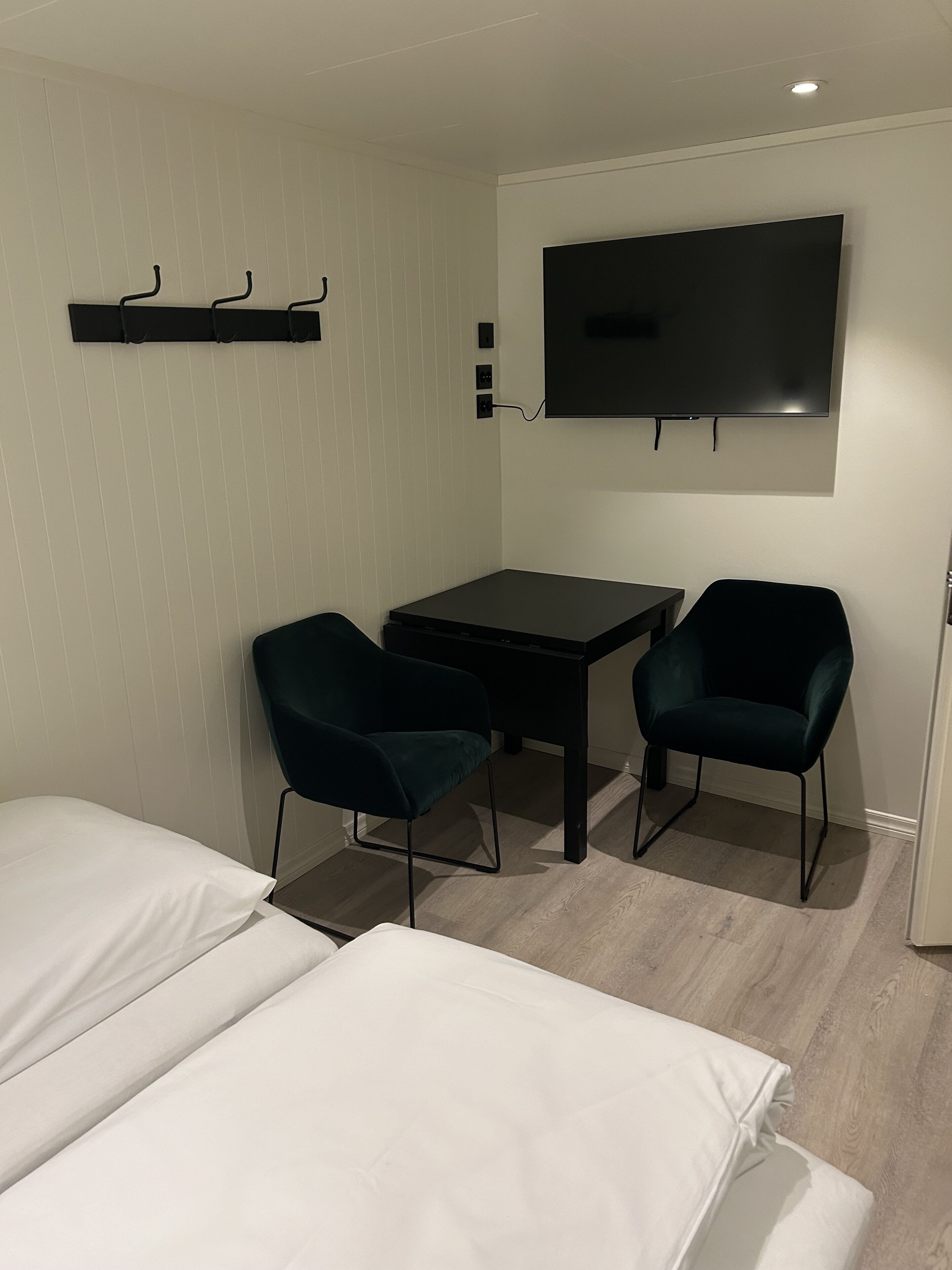 A small, modern room with a bed, a desk, two chairs, and a TV mounted on the wall.