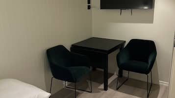 A small, modern room with a bed, a desk, two chairs, and a TV mounted on the wall.