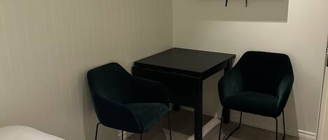 A small, modern room with a bed, a desk, two chairs, and a TV mounted on the wall.