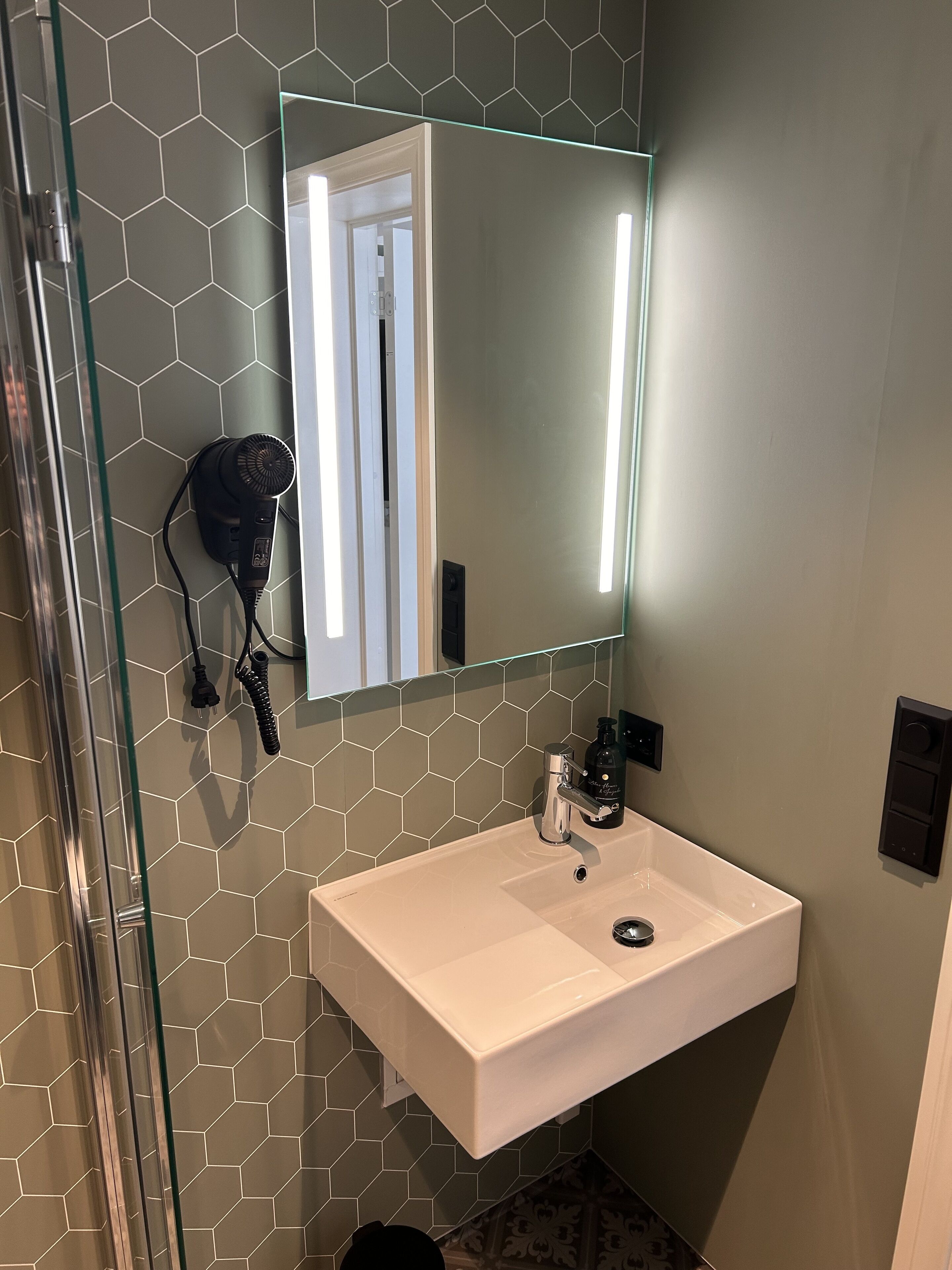 A bathroom with a white sink, a mirror with a light, and a hairdryer on the wall.