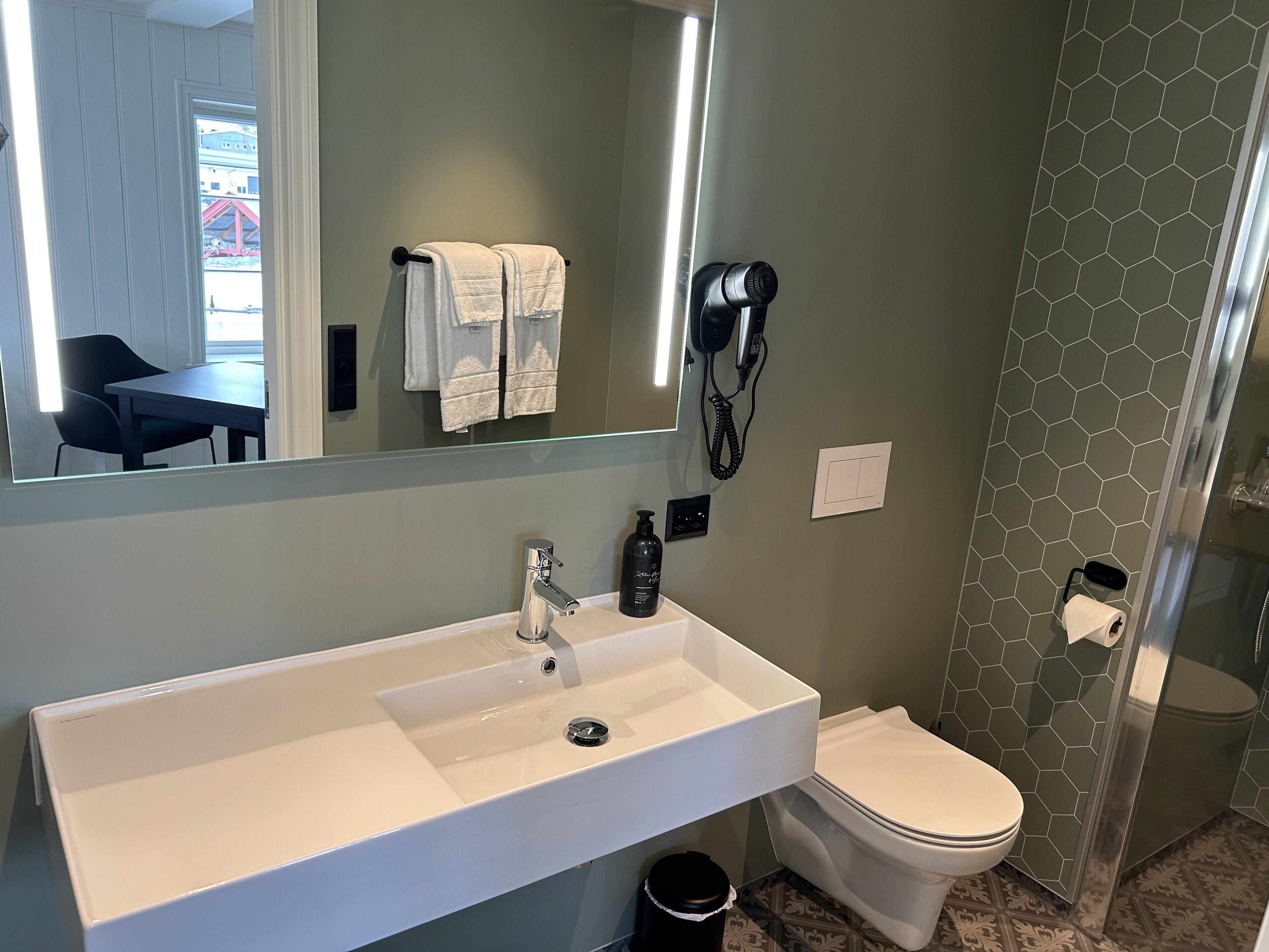 Une salle de bain avec un lavabo blanc, des toilettes, un sèche-cheveux fixé au mur et un miroir au-dessus du lavabo.