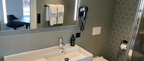 Une salle de bain avec un lavabo blanc, des toilettes, un sèche-cheveux fixé au mur et un miroir au-dessus du lavabo.