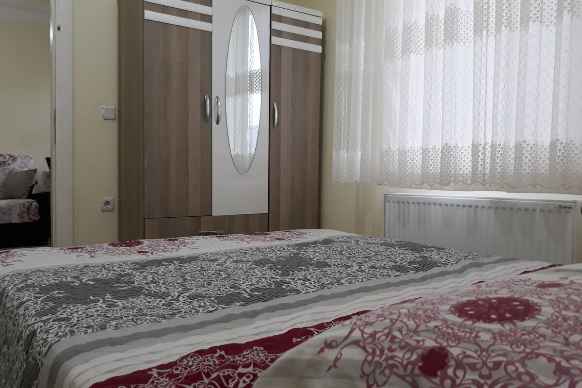 Quarto com uma cama, um guarda-roupa, um radiador e uma janela com cortinas leves.