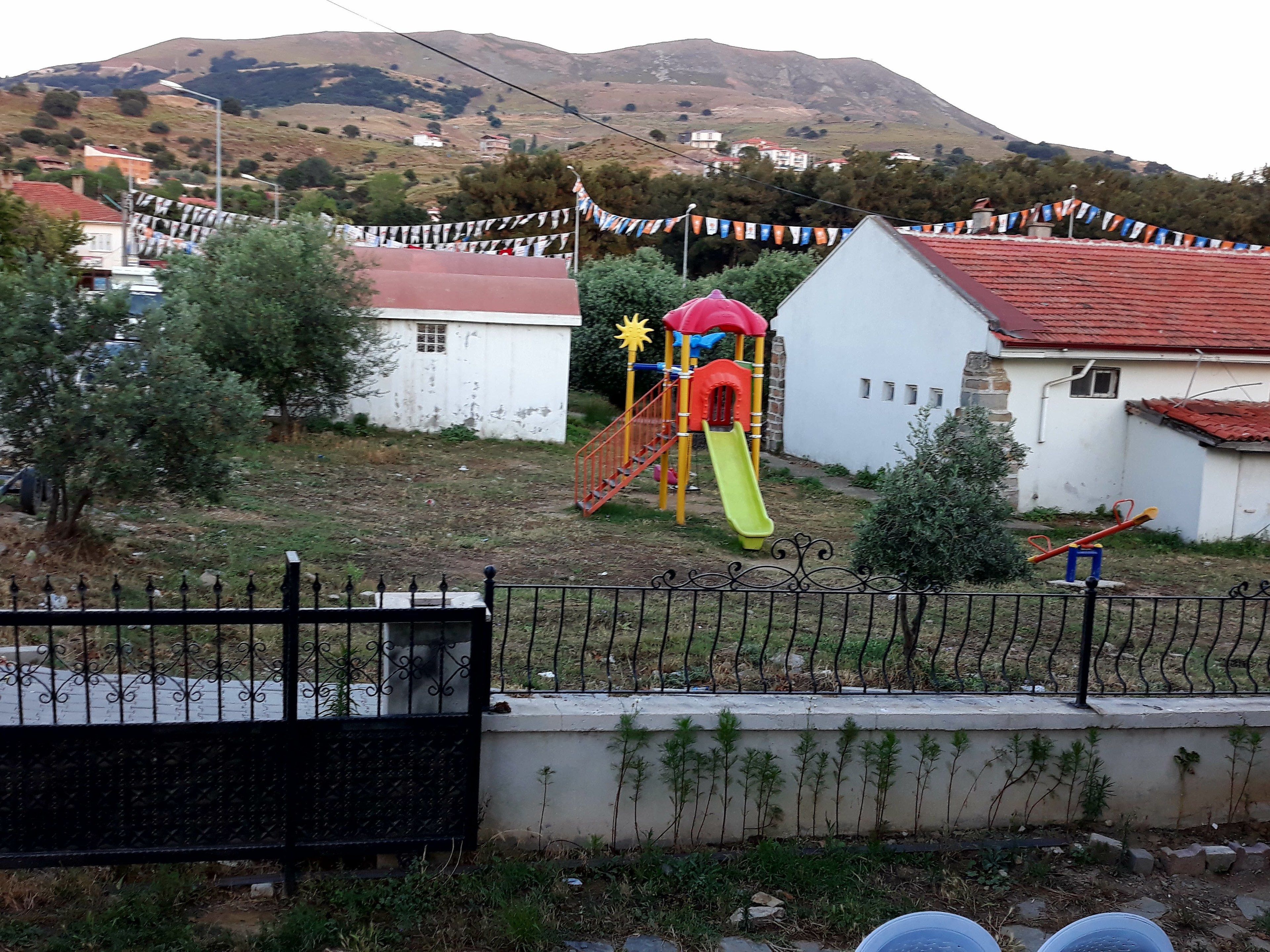A playground with a slide and climbing structures, surrounded by a fence and houses in a hilly area.