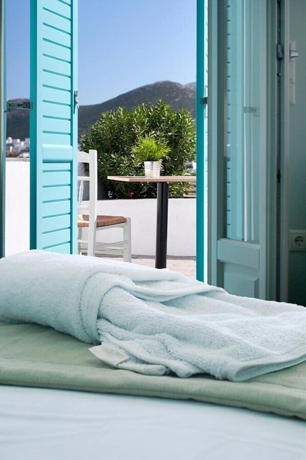 A bed with a white blanket, a chair, a small table with a plant, and a view of mountains through open blue shutters.