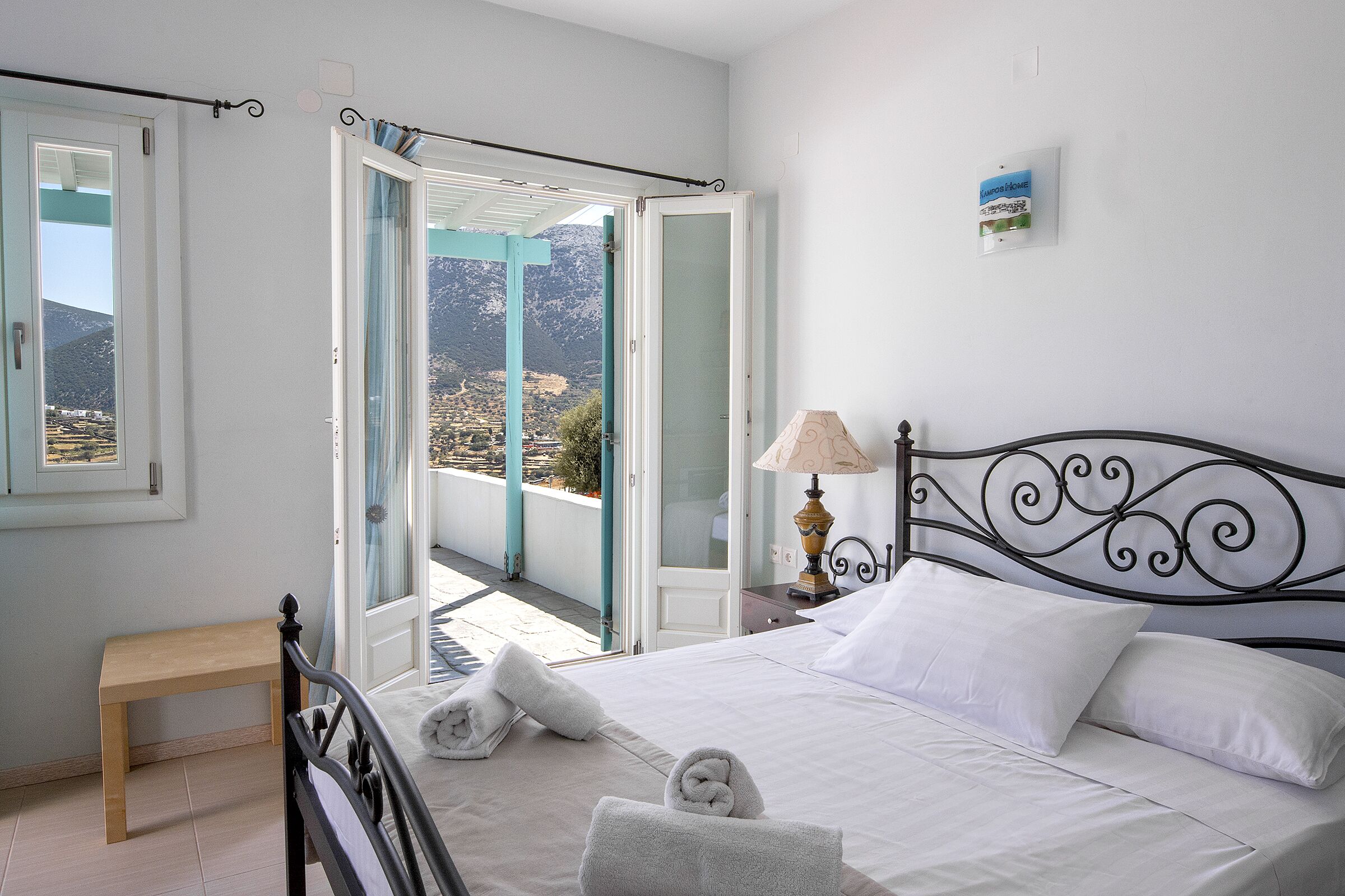 Un dormitorio con cama, mesita de noche, lámpara y puerta abierta que da a un balcón con vistas a montañas.