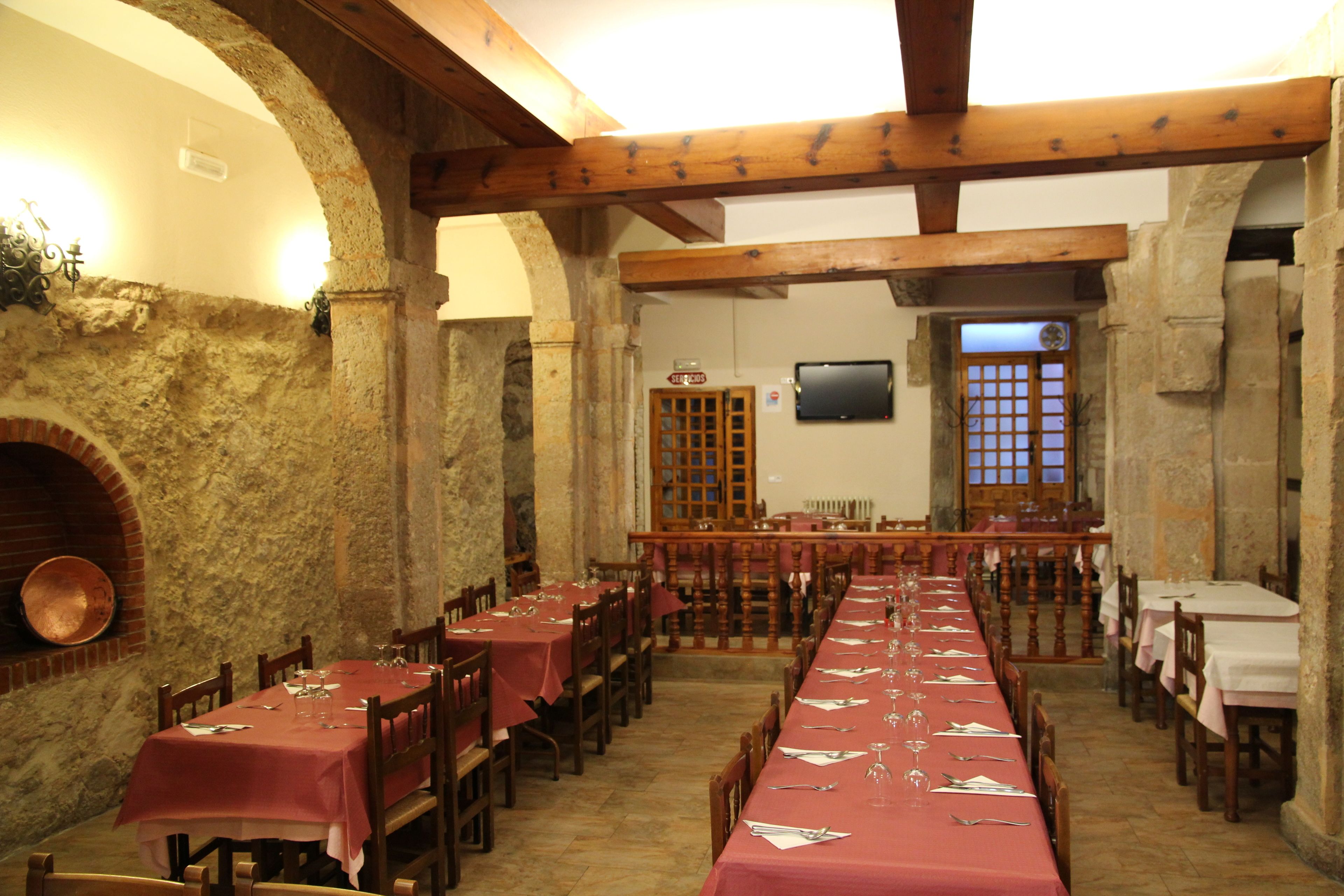 A long dining table with red tablecloths and wooden chairs, set in a stone-walled room with a fireplace and wooden beams.