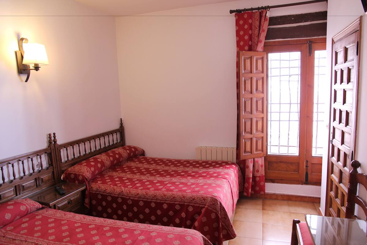 Un dormitorio con tres camas, cabecera de madera, mesita de noche, una mesa y una ventana con puerta de madera.