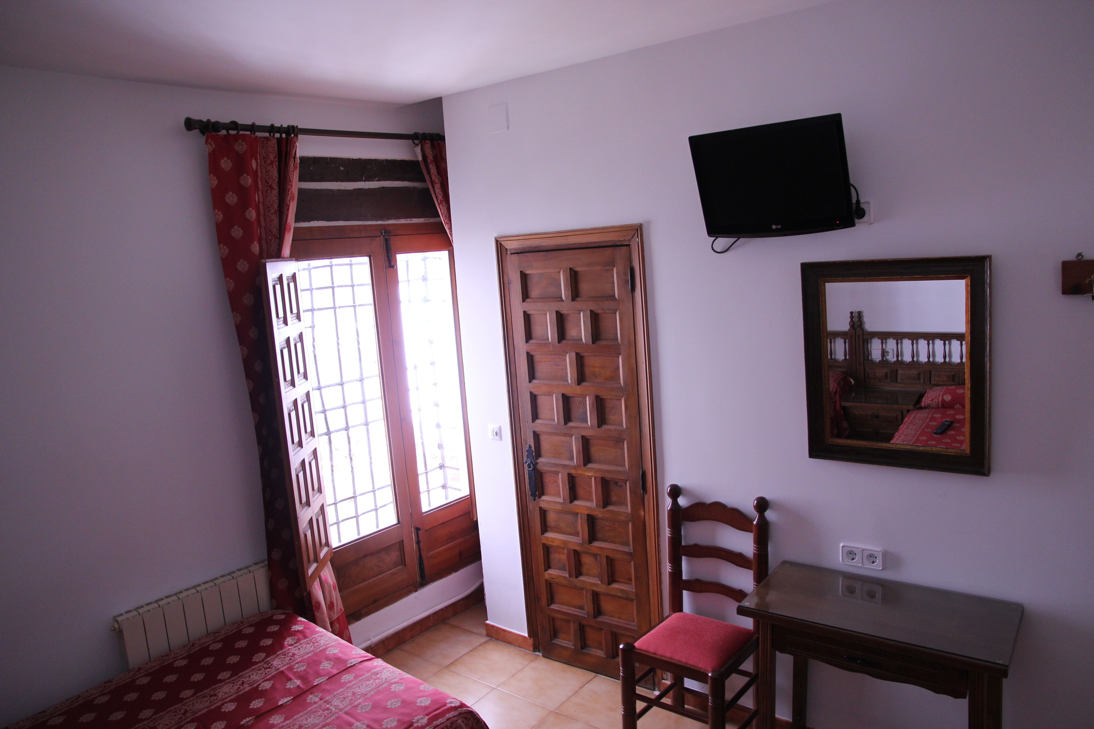 Un dormitorio con una cama, una puerta de madera, una silla, un escritorio, un televisor y un espejo.