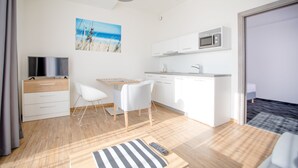 Una habitación moderna y luminosa con suelo de madera, un comedor con sillas blancas, una cocina con armarios blancos y un microondas, y un televisor sobre un mueble de madera.