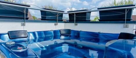 A blue hot tub with a glass enclosure, surrounded by a white wall and a clear blue sky with clouds.