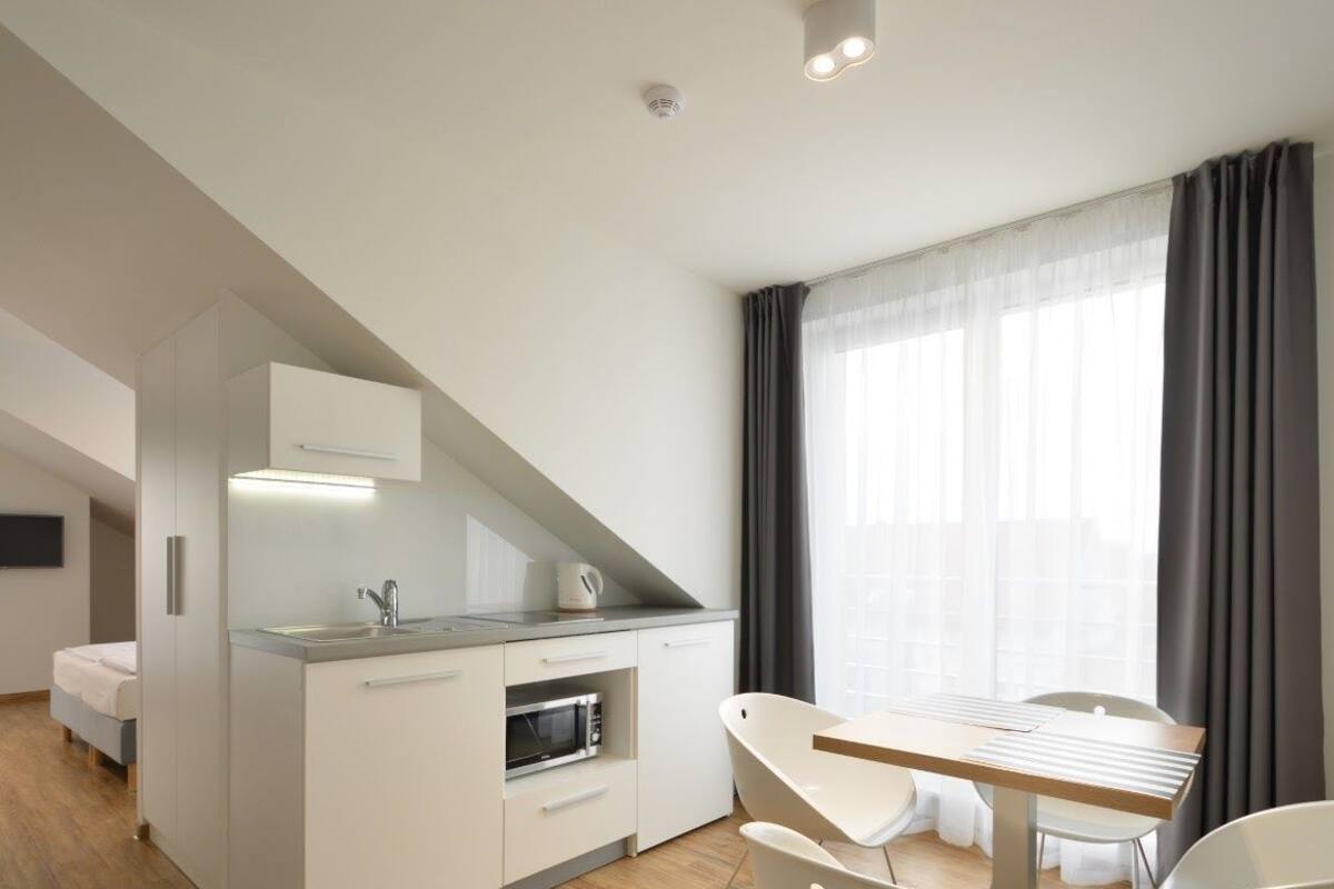A modern hotel room with a kitchenette, a bed, a TV, and a large window with curtains.