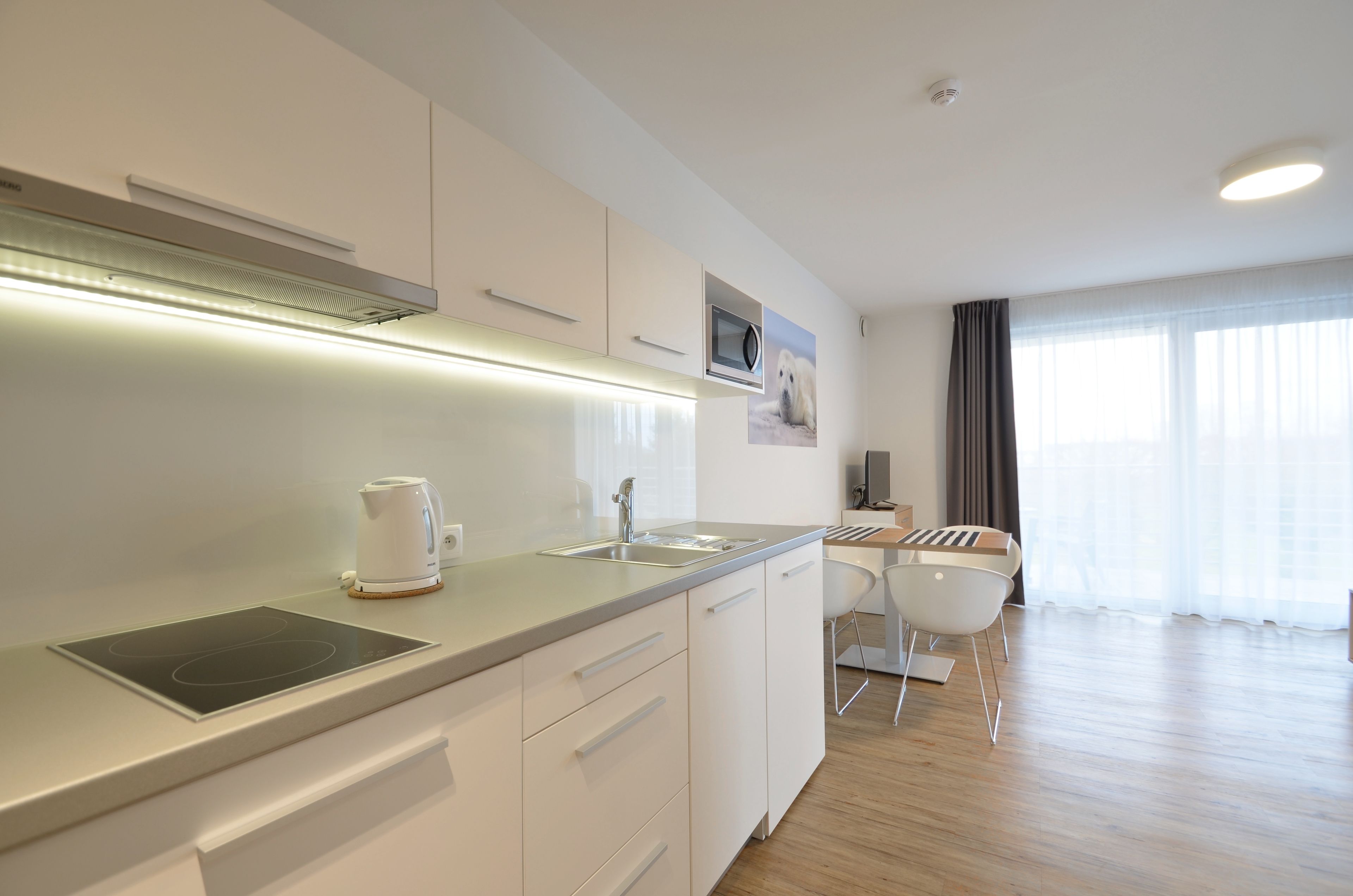 A modern kitchen with a built-in stove, a kettle, and a sink. There is a dining area with a table and chairs.